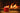 A mug of hot tea with a sweater tea cozy on the mug, on a wooden table with a fire in the background.