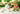 A bright garden picture of a tea party - a white porcelain teapot, a cup  of tea, and a gold infuser ball with a pitcher of cream, plus a three-tier tray of sandwiches, tarts, and other afternoon tea pastries.