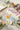 A styled flat-lay of colorful felt tea bag charms shaped like teacups and teacups with floral prints, arranged on a light fabric surface with tea cups, flowers, books, and a teapot.