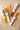 Four boxes of honey sticks arranged on a light beige stone surface, with loose strands of yellow and orange pasta fanned out beside them. The boxes are white with small colored labels and a honeycomb logo.