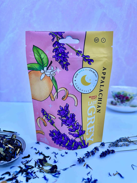 A pouch of Appalachian Lady Grey Tea with lavender buds, cornflowers, and tea spilling out of a tea infuser with a china tea cup in the background.