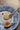 A round baked cookie sits on a white lace doily on a blue-and-white patterned plate, with a silver fork resting beside it and a halved fig and a small red berry on the plate, all set on a wooden surface.