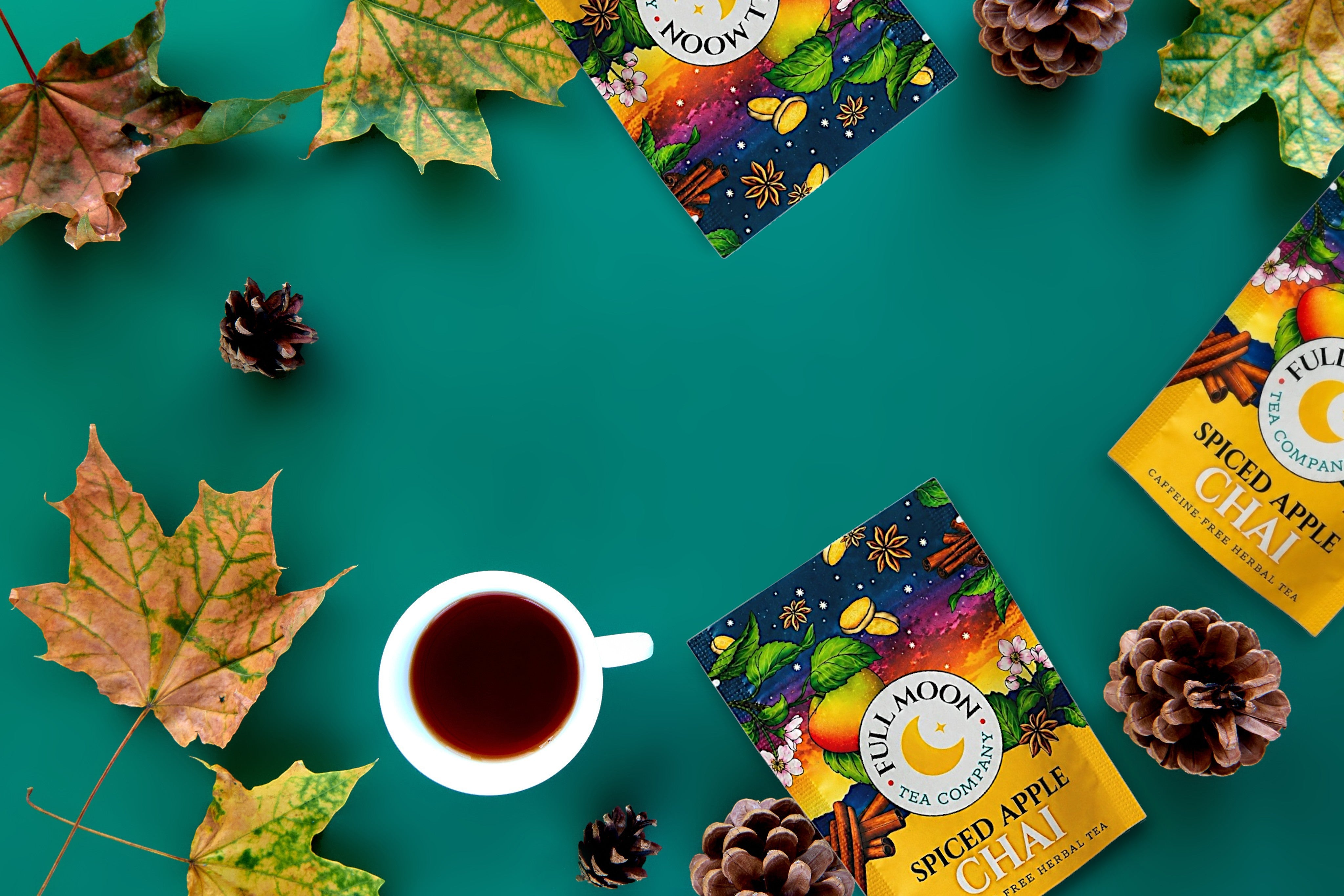 Three boxes of Full Moon Extra Apple Chai tea arranged on a teal background with autumn maple leaves, pinecones, and a white cup of tea.