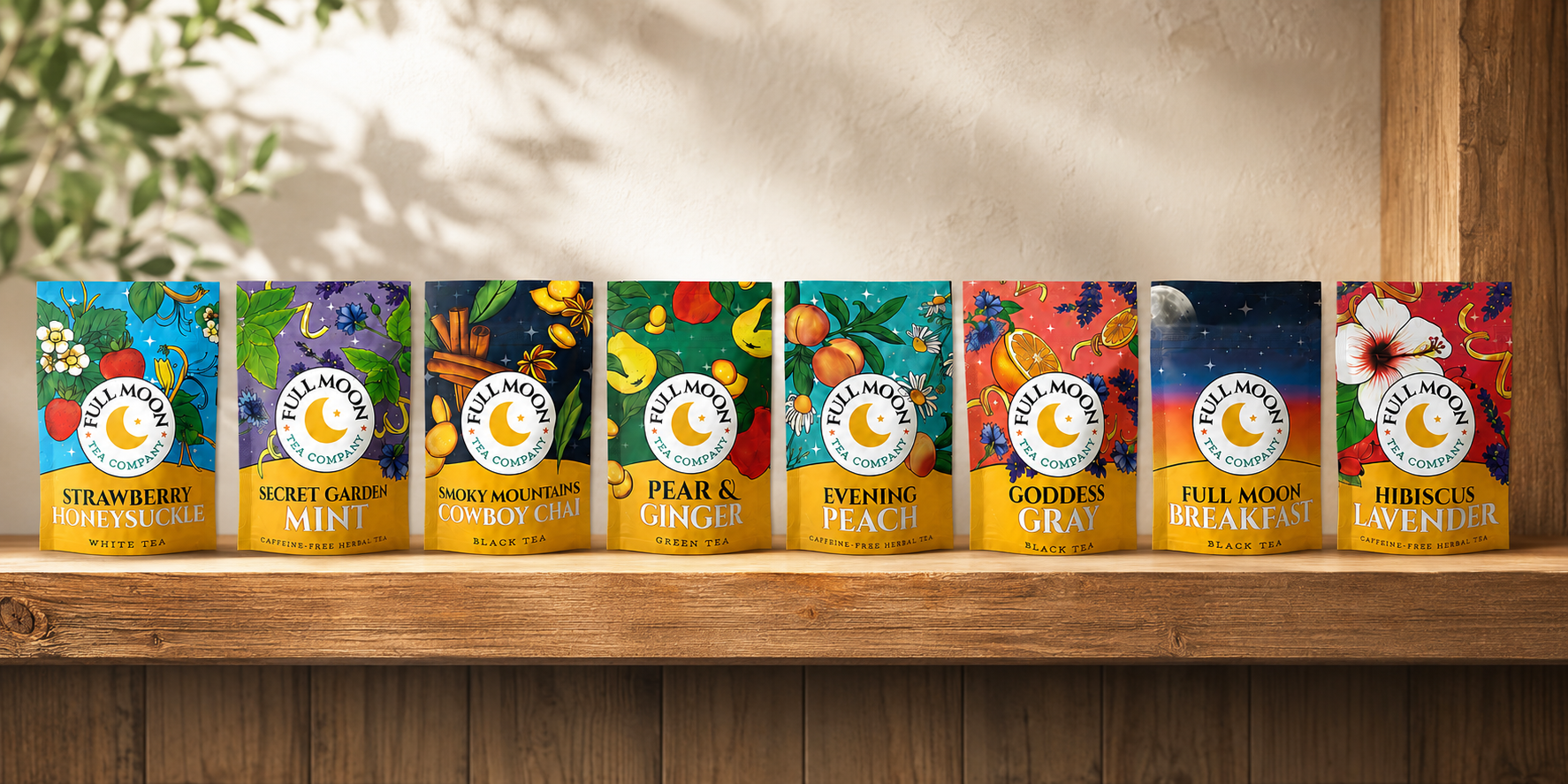 Eight colorful tea tins from Tea Moon Tea Company arranged in a row on a wooden shelf. The labels show different tea flavors: Strawberry Breakfast, Suede Moon Mint, Sun Moon Cowboy Coffee, Ginger, Pear & Peach, Goddess Gray, Full Moon Breakfast, Full Moon Lunch, Full Moon Dinner, and Full Moon Awener.