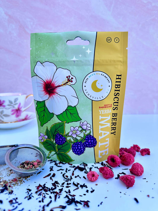  A colorful pouch of Hibiscus Berry Yerba Mate organic tea from Full Moon Tea Company on a soft pink background with a teacup, loose leaf tea, and dried raspberries and hibiscus around it.