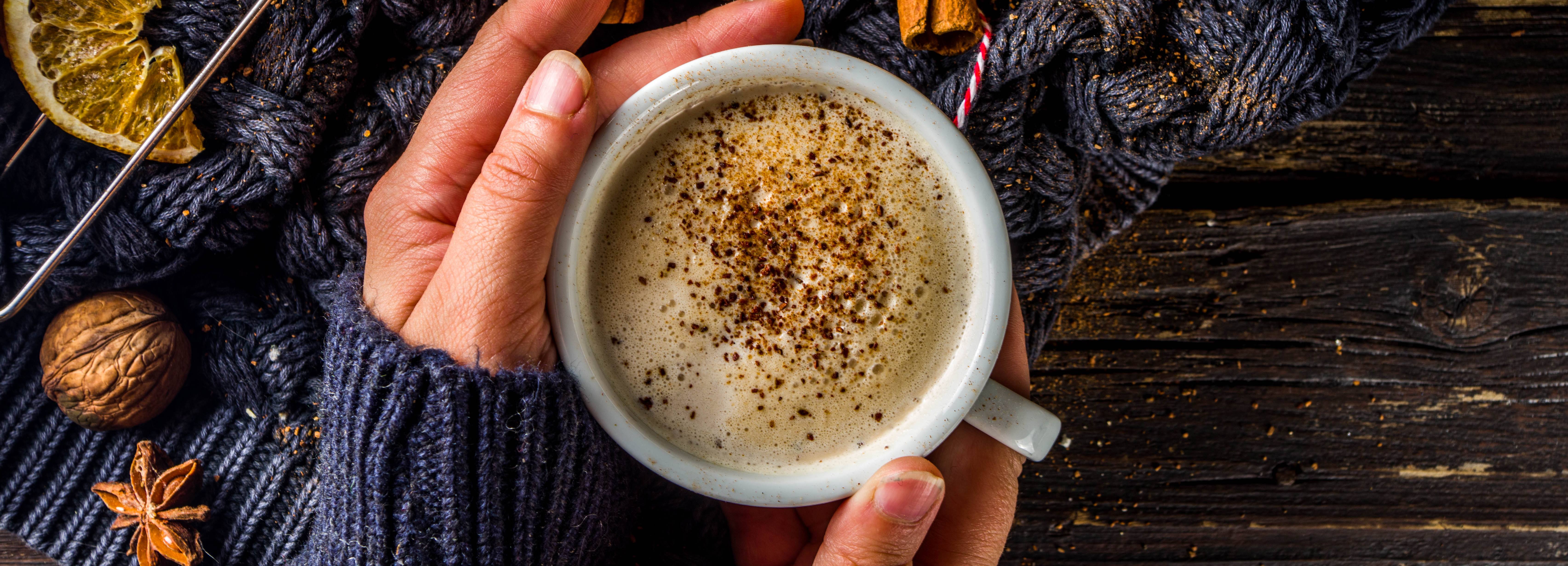 Person holding a mug of hot chocolate with whipped cream and nutmeg on a wooden surface.