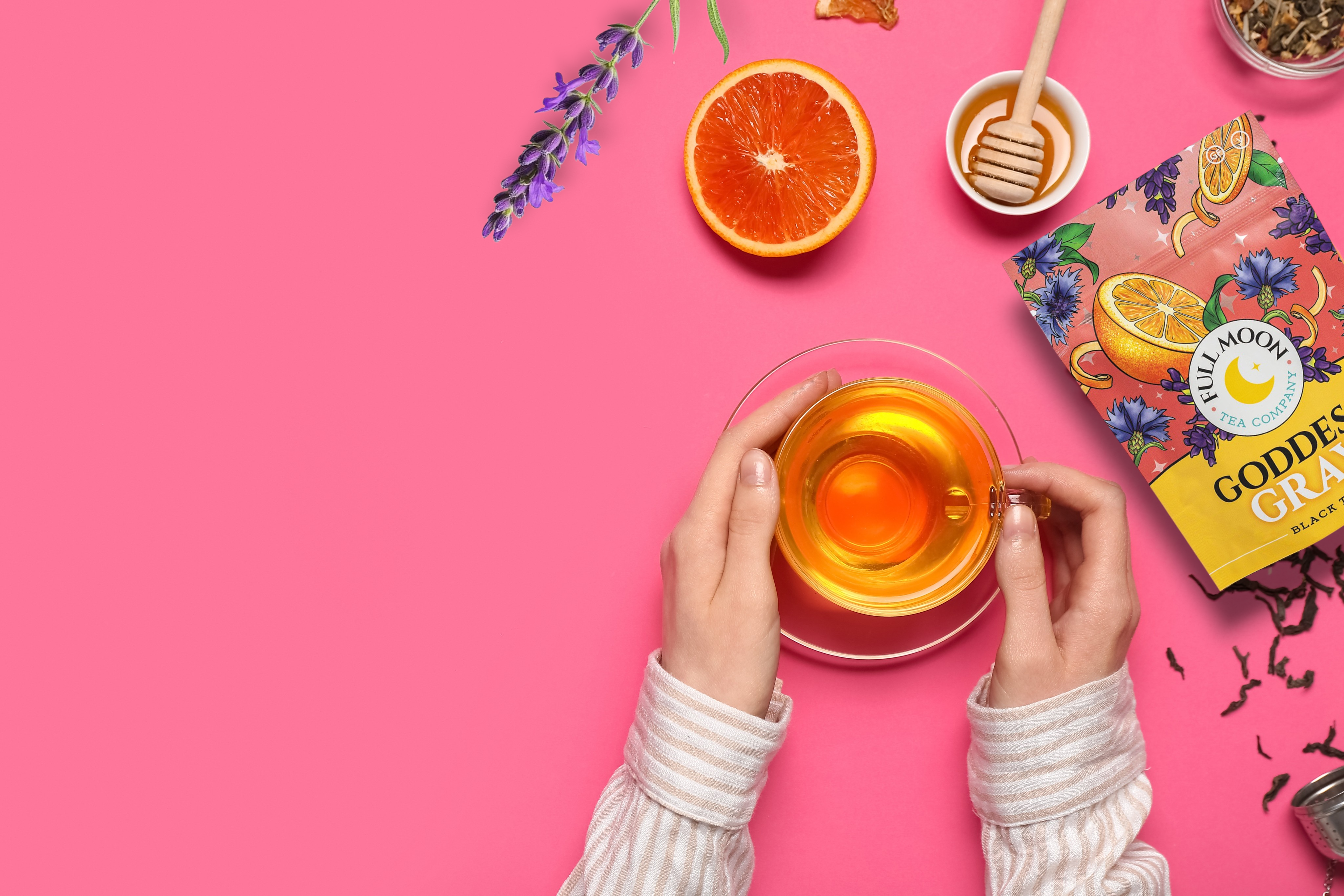 Person holding a glass of tea with a pink background, featuring a tea box and tea leaves.