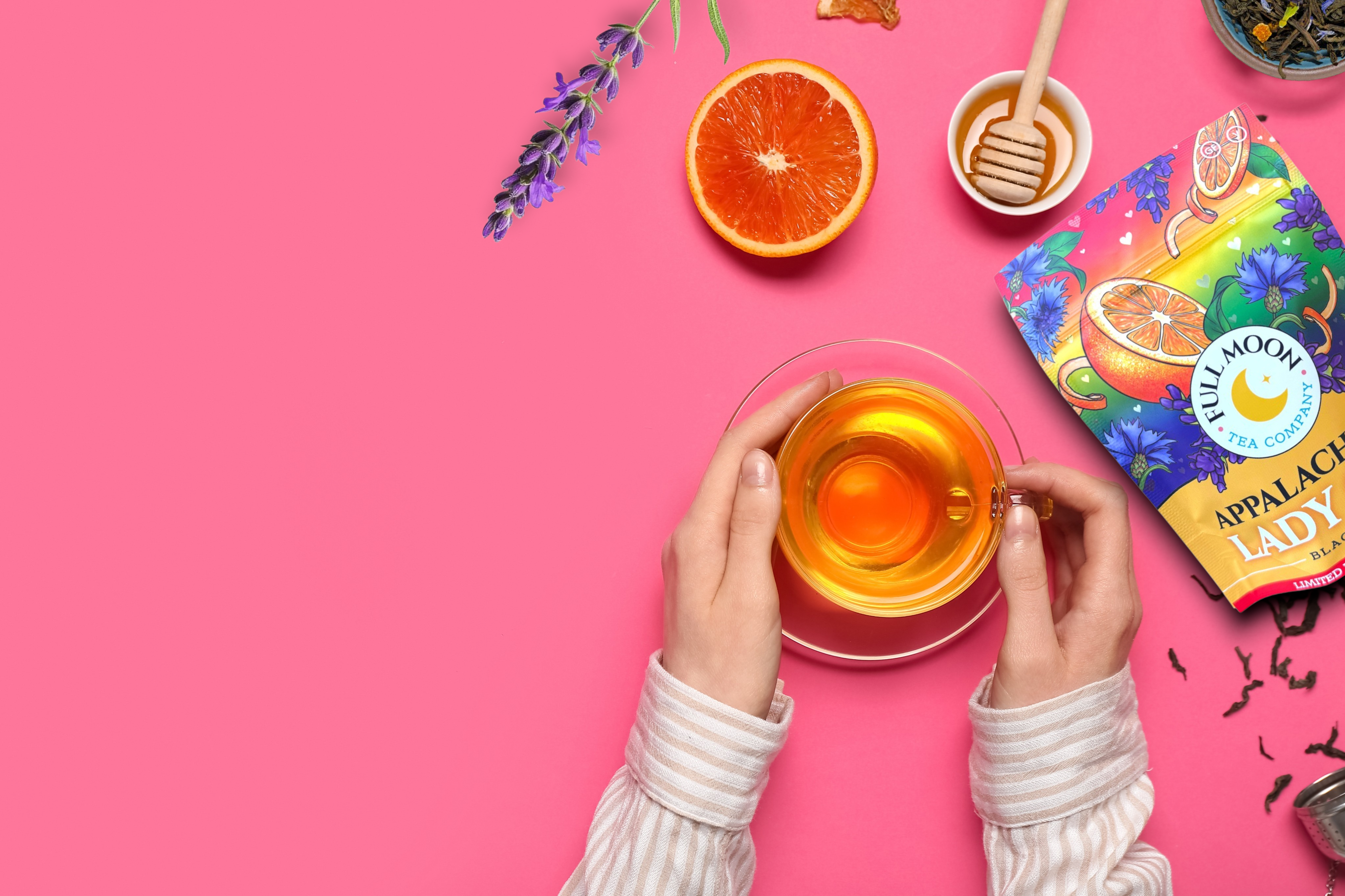 Person holding a glass of tea with a pink background, featuring a tea bag and fruits.