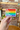 Hand holding a pride rainbow sticker with the words be gay drink tea fight fascism against a brick background