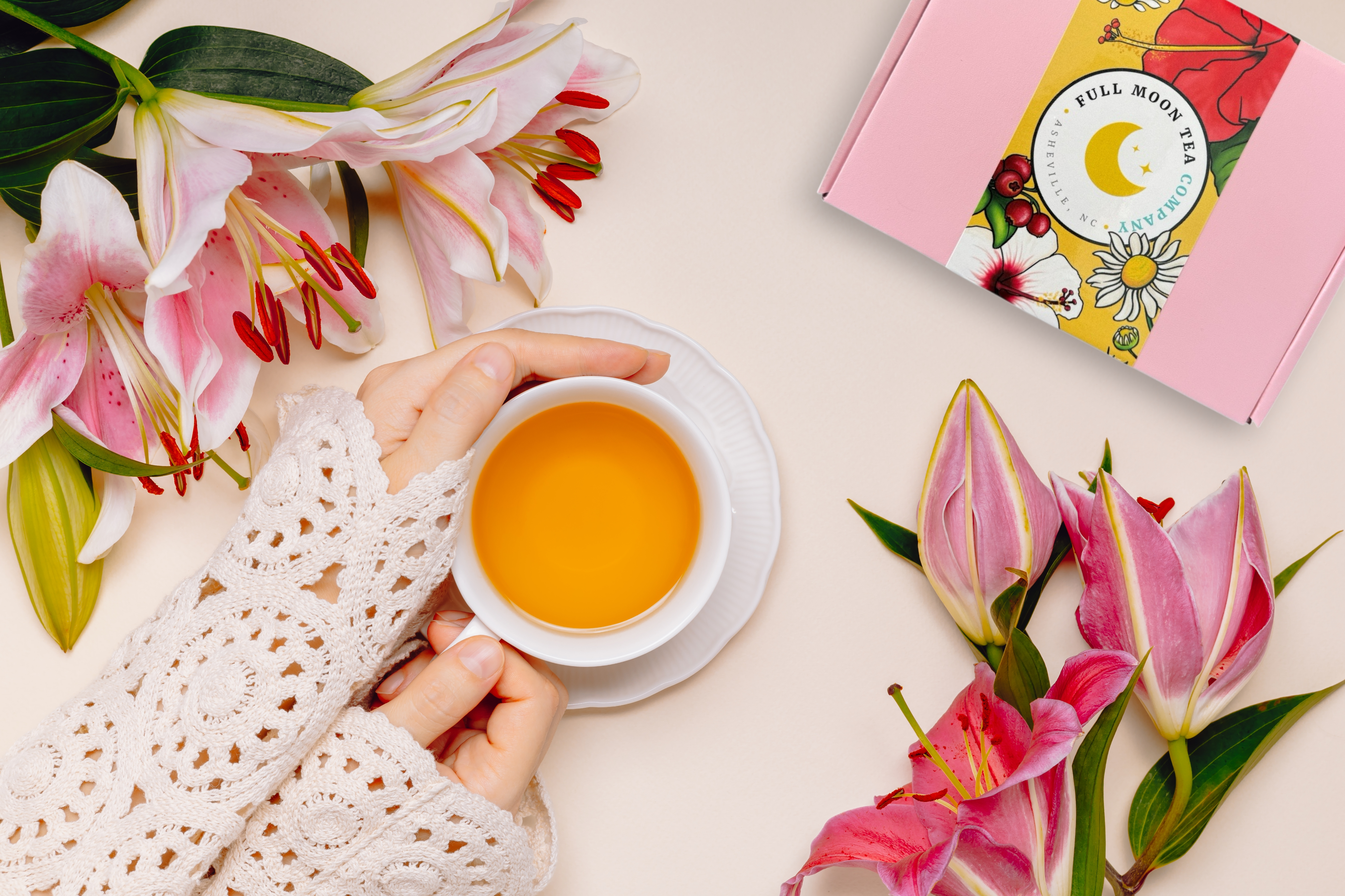 Person holding a cup of tea with pink lilies and a tea box in the background
