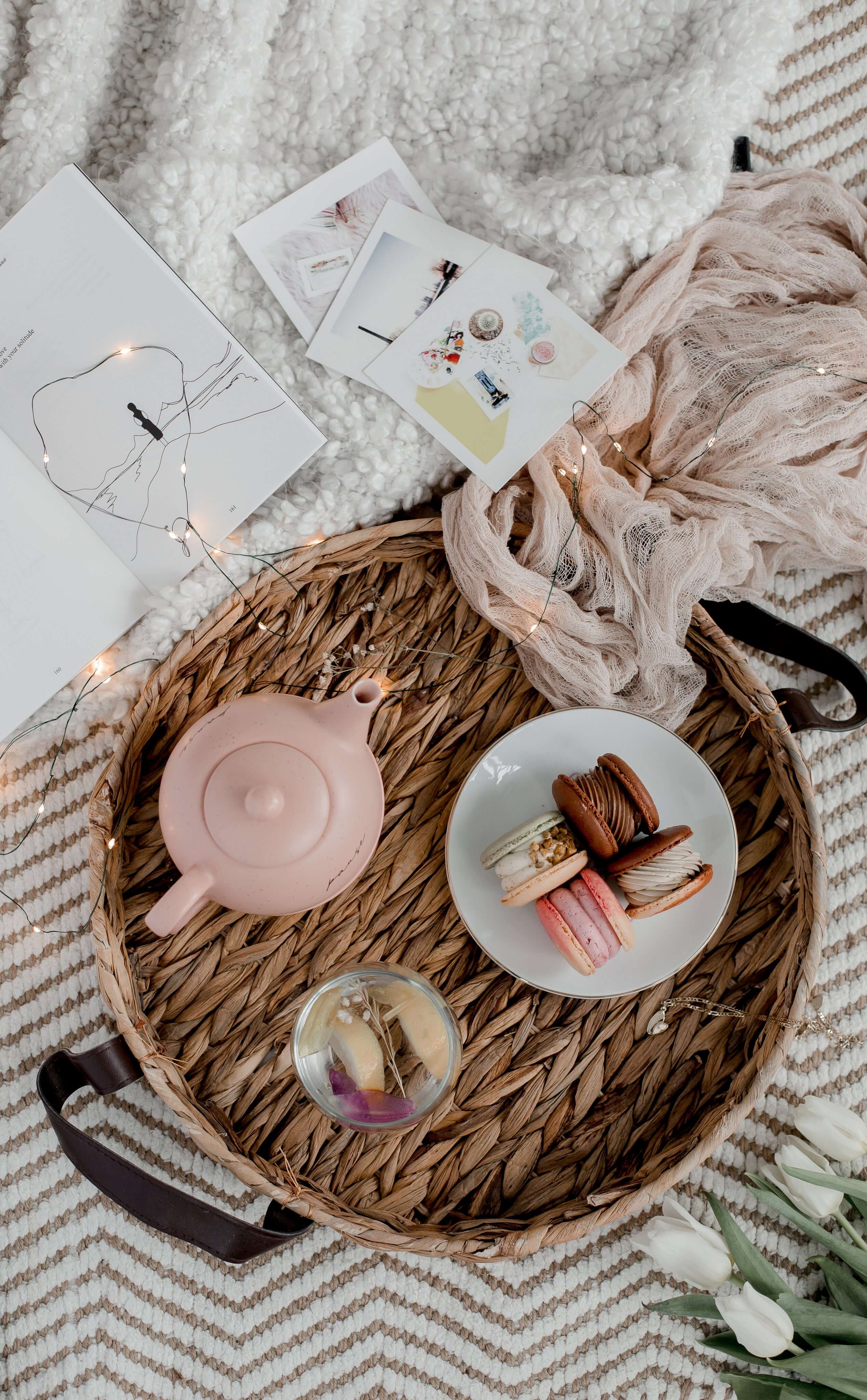 A whicker tray holding a pink teapot, a cup of floral tea and a plate of macarons on a textile background with a soft, organic scarf and flowers.